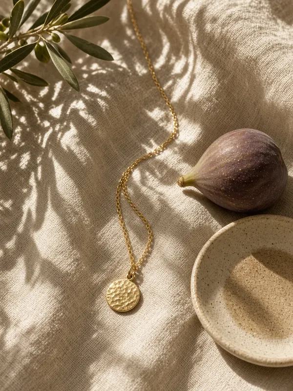 Gold chain necklace with a hammered disc on linen with olive leaf shadows and a fig